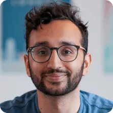 Smiling young man with dark curly hair, glasses, and a beard wearing a blue shirt.